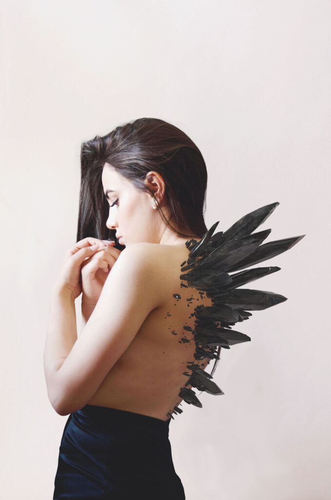 Valeria Cisneros portrays a poised introspective figure with a striking dark crystalline structure emerging from her back set against a soft neutral background.
